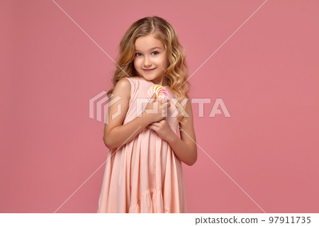 Little girl with a blond curly hair, in a pink dress is posing with a candy 97911735