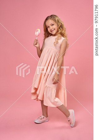 Little girl with a blond curly hair, in a pink dress is posing with a candy Little girl with a blond curly hair, in a pink dress is posing with a candy 97911796