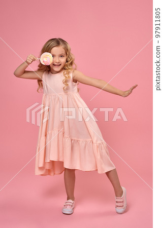 Little girl with a blond curly hair, in a pink dress is posing with a candy 97911805