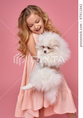 Little girl with a blond curly hair, in a pink dress plays with her dog Little girl with a blond curly hair, in a pink dress plays with her dog 97911842