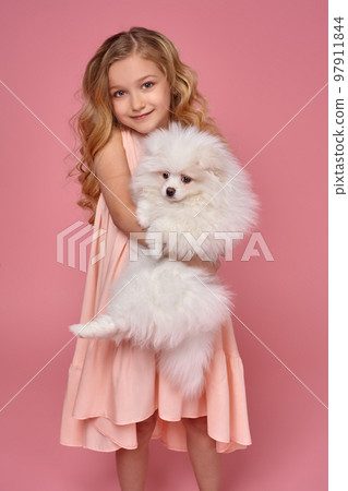 Little girl with a blond curly hair, in a pink dress plays with her dog Little girl with a blond curly hair, in a pink dress plays with her dog 97911844