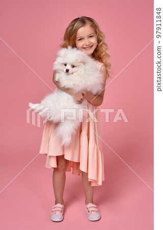 Little girl with a blond curly hair, in a pink dress plays with her dog Little girl with a blond curly hair, in a pink dress plays with her dog 97911848