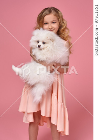 Little girl with a blond curly hair, in a pink dress plays with her dog 97911851