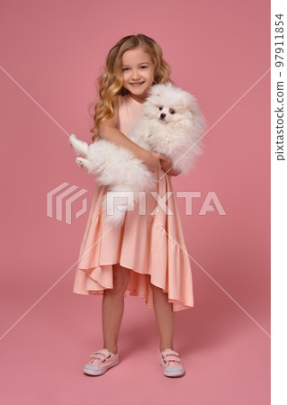 Little girl with a blond curly hair, in a pink dress plays with her dog Little girl with a blond curly hair, in a pink dress plays with her dog 97911854