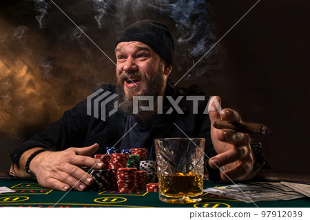 Man is playing poker with a cigar and a whiskey. A man winning all the chips on the table with thick cigarette smoke. Man is playing poker with a cigar and a whiskey. A man winning all the chips on the table with thick cigarette smoke. 97912039