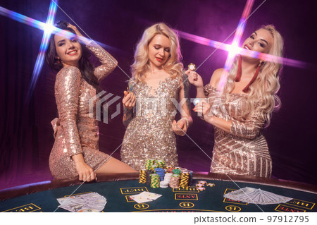 Beautiful girls with a perfect hairstyles and bright make-up are posing standing at a gambling table. Casino, poker. 97912795