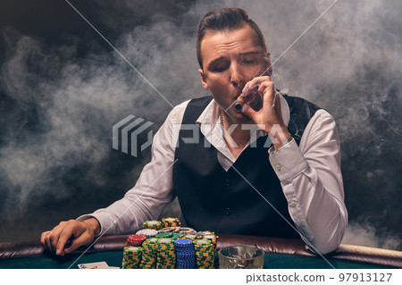 Wealthy man is smoking a cigar and playing poker with an excitement at a casino on black background. 97913127