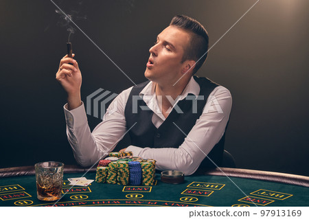 Wealthy man is smoking a cigar and playing poker with an excitement at a casino on black background. 97913169
