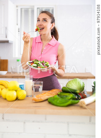 Slim young woman eating vegetable salad in home kitchen Slim young woman eating vegetable salad in home kitchen 97914108