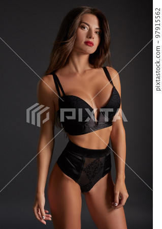 Stunning caucasian female model with dark hair and red lips in black underwear posing on dark grey background. 97915562