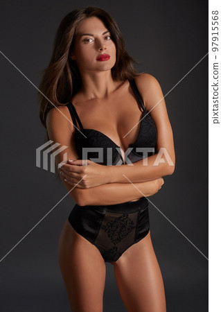 Stunning caucasian female model with dark hair and red lips in black underwear posing on dark grey background. 97915568