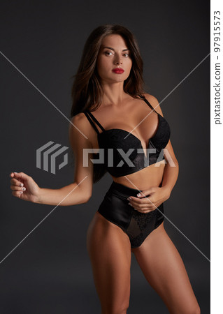 Stunning caucasian female model with dark hair and red lips in black underwear posing on dark grey background. Stunning caucasian female model with dark hair and red lips in black underwear posing on dark grey background. 97915573