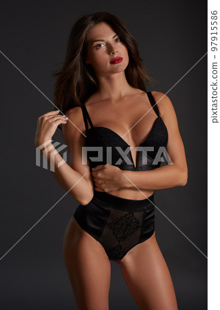 Stunning caucasian female model with dark hair and red lips in black underwear posing on dark grey background. 97915586