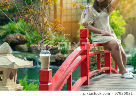 Iced cappuccino with straws placed on head of red wooden bridge with blurred image of woman sit on bridge railing and small waterfalls and cool flowing pond in garden decorated in Japanese style. Iced cappuccino with straws placed on head of red wooden bridge with blurred image of woman sit on bridge railing and small waterfalls and cool flowing pond in garden decorated in Japanese style. 97920081