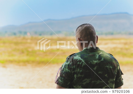 Back view of Asian army soldier stands to explore the landscape in field. Command class soldier stands to watch and inspect the conditions of the maneuver in open terrain with mountain in background. 97920106