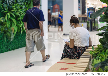 Back view of Asian woman wear face mask to prevent Corona virus (Covid-19) sit and using a smart phone and small notebook in public place and keep social distancing with other people. New normal. 97920357