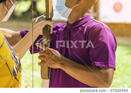 Young woman practice archery with male instructor closely supervising. Young female archer, young female archer, archery, shoot arrow with bow to target, archery training. Selective focus on hands. 97920489