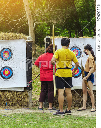 Back view of Asian archers wear face mask collect arrows fired from a bow on sport target. Archer clearing arrows from target to start new game. Meditation exercise from archery and outdoor recreation Back view of Asian archers wear face mask collect arrows fired from a bow on sport target. Archer clearing arrows from target to start new game. Meditation exercise from archery and outdoor recreation 97920526