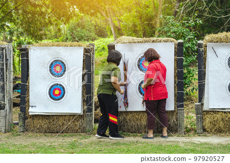 Back view of female officer wear face mask collect arrows fired from a bow on sport target. Women clearing arrows from target to start new game. Meditation exercise from archery and outdoor recreation Back view of female officer wear face mask collect arrows fired from a bow on sport target. Women clearing arrows from target to start new game. Meditation exercise from archery and outdoor recreation 97920527