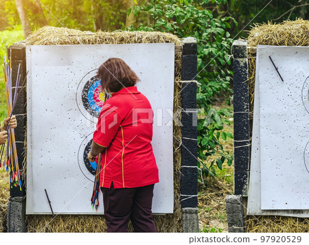 Back view of female officer wear face mask collect arrows fired from a bow on sport target. Woman clearing arrows from target to start new game. Meditation exercise from archery and outdoor recreation Back view of female officer wear face mask collect arrows fired from a bow on sport target. Woman clearing arrows from target to start new game. Meditation exercise from archery and outdoor recreation 97920529