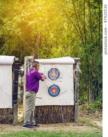 Back view of male officer wear face mask collect arrows fired from a bow on sport target. Archer clearing arrows from target to start new game. Meditation exercise from archery and outdoor recreation. Back view of male officer wear face mask collect arrows fired from a bow on sport target. Archer clearing arrows from target to start new game. Meditation exercise from archery and outdoor recreation. 97920530