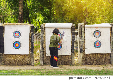 Back view of female officer wear face mask collect arrows fired from a bow on sport target. Woman clearing arrows from target to start new game. Meditation exercise from archery and outdoor recreation 97920531