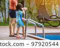 Adorable daughter in blue swimming suit going down swimming pool ladder to swim with clear blue water and her mother to take care of closely. Little girl at pool. Happy family relationship at pool. 97920544