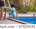 Adorable daughter in blue swimming suit going down swimming pool ladder to swim with clear blue water and her mother to take care of closely. Little girl at pool. Happy family relationship at pool. 97920545