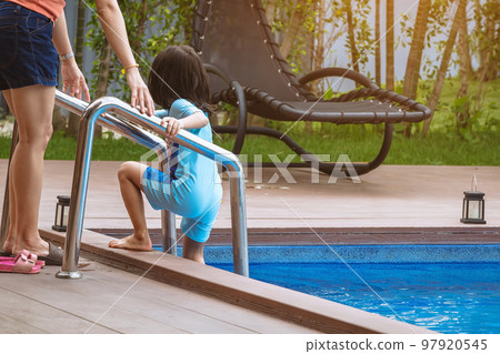 Adorable daughter in blue swimming suit going down swimming pool ladder to swim with clear blue water and her mother to take care of closely. Little girl at pool. Happy family relationship at pool. Adorable daughter in blue swimming suit going down swimming pool ladder to swim with clear blue water and her mother to take care of closely. Little girl at pool. Happy family relationship at pool. 97920545