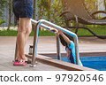 Mother wear short jeans take care her lovely daughter in blue swimming suit going down swimming pool ladder to swim with clear blue water. Mom and daughter at pool. Happy family relationship at pool. 97920546