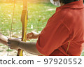 Female teacher teaches student to aim at goal. An archer teaching young man archery on field. Instructor teaching man to use bow and arrow on archery training. Archery course. Selective focus on hand. 97920552