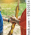 Female teacher teaches student to aim at goal. An archer teaching young man archery on field. Instructor teaching man to use bow and arrow on archery training. Archery course. Selective focus on hand. 97920554