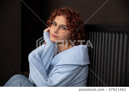 A girl with curly hair and glasses sits by a hot radiator. A girl with curly hair and glasses sits by a hot radiator. 97921482