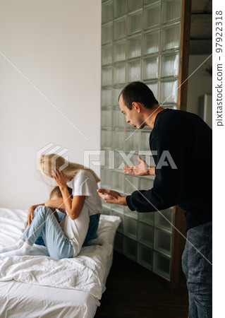Vertical shot of scared mother and daughter hugging each other sitting on sofa while aggressive father angrily yelling at them at home. Drunken husband scolding, raising voice, scream at family 97922318