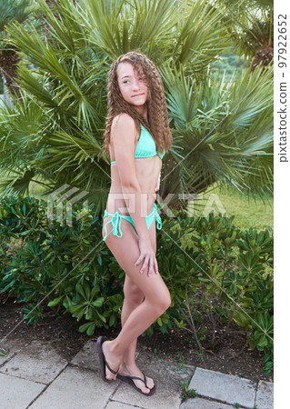 Teenage girl with freckles and curly long hair in bathing suit poses against backdrop of palm tree 97922652