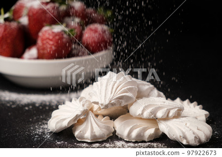 Powdered sugar falls on meringue and strawberries on a black background. 97922673