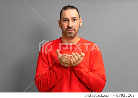 Latino man with beard dressed in a red jumper offering something imaginary in his hands close to the chest isolated on gray studio background, advertising concept, copy space 97923262