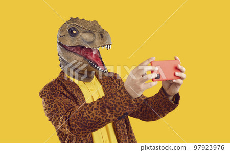 Eccentric funny man in rubber dinosaur mask taking selfie photo on smartphone on yellow background. 97923976