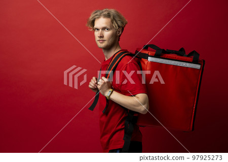 Portrait of young courier with curly hair in red uniform with red thermo bag on red background looks straight in camera. Food delivery service. 97925273