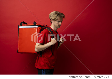 Side view of young courier with curly hair in red uniform with red thermo bag on red background. Food delivery service. Side view of young courier with curly hair in red uniform with red thermo bag on red background. Food delivery service. 97925274