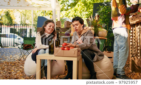 Woman sitting at table with winemaker tasting homemade organic wine, enjoying product sampling before purchasing produce. Shopping at natural street food festival farmers market. Woman sitting at table with winemaker tasting homemade organic wine, enjoying product sampling before purchasing produce. Shopping at natural street food festival farmers market. 97925957