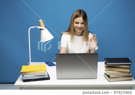 Front view young female freelancer sitting at studio before laptop screen waving hand to web camera. Student starting distant lesson, lecture or training course online. Front view young female freelancer sitting at studio before laptop screen waving hand to web camera. Student starting distant lesson, lecture or training course online. 97925968