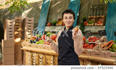 Farmers market vendor showing organic produce to clients, presenting colorful fruits and vegetables on food marketplace stand. Young adult having farming counter to sell products. 97925990