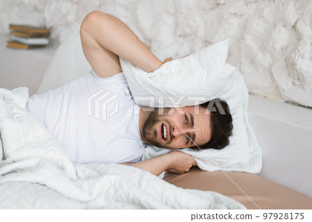 Tired angry sad young caucasian guy lies on bed, covers his ears with pillow, suffers from noisy neighbors 97928175