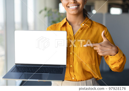Female black entrepreneur pointing at laptop computer with blank screen for website design, standing in office, mockup 97928176