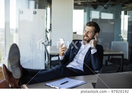 Happy middle aged businessman relaxing in office, resting feet on table using cellphone and listening to music 97928192