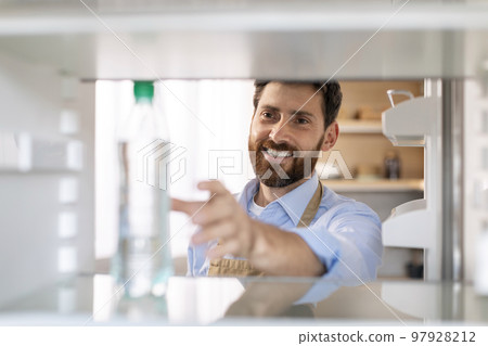 Smiling millennial caucasian bearded guy in apron opens door of empty refrigerator and takes bottle of water 97928212