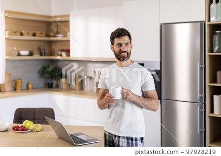Happy millennial caucasian guy with beard with cup of coffee enjoys good morning, has breakfast with computer 97928219