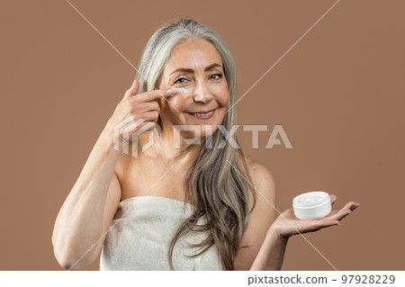Smiling mature caucasian woman with natural beauty shows jar and applies cream on face, recommend rejuvenation 97928229