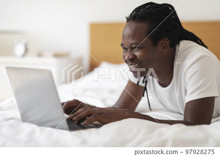 Online Communication. Smiling African American Guy Using Laptop While Relaxing In Bed Online Communication. Smiling African American Guy Using Laptop While Relaxing In Bed 97928275